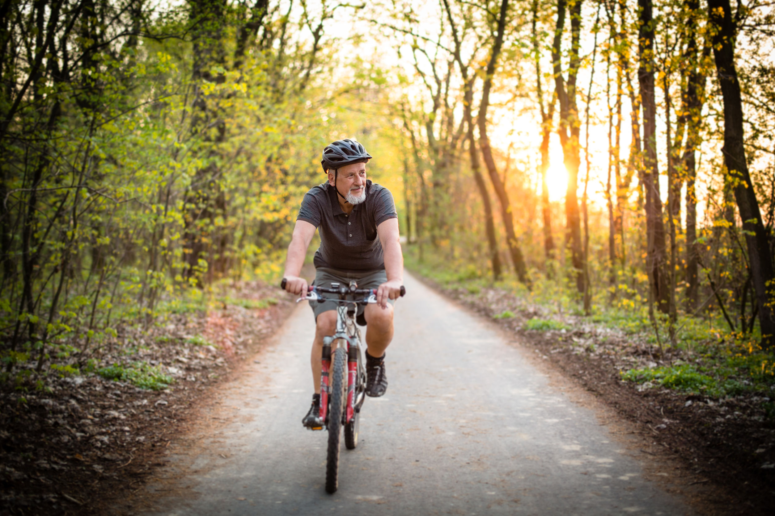 Ein älterer Mann auf einem Mountainbike fahrt auf einem Radweg durch einen Wald. Im Hintergrund geht die Sonne unter. Der Mann trägt ein T-Shirt, Helm und eine kurze Hose.